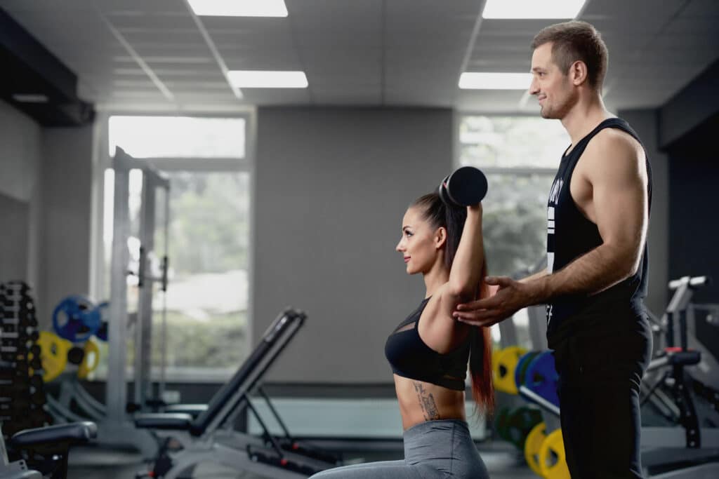 Male personal trainer helping woman working with heavy dumbbells at gym.